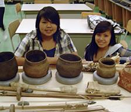 Vase reproductions and other Algonquin objects on a table. Two teenagers looking at the photographer in the background.