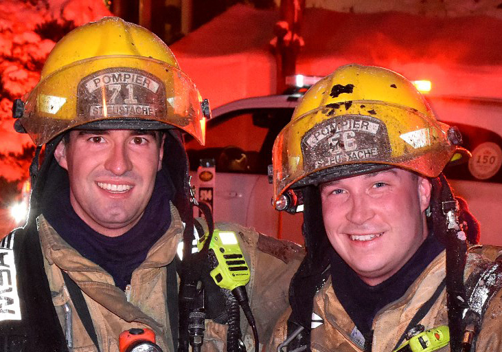 Photograph of two smiling firefighters in firefighting gear, in front of a car, a fire truck and many houses, at night, under the lights.