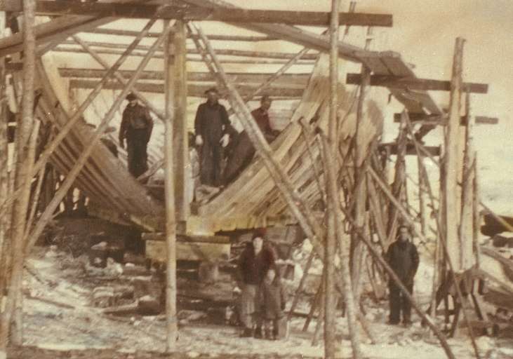 View of a schooner under construction. The structure of the ship is surrounded by scaffolding.