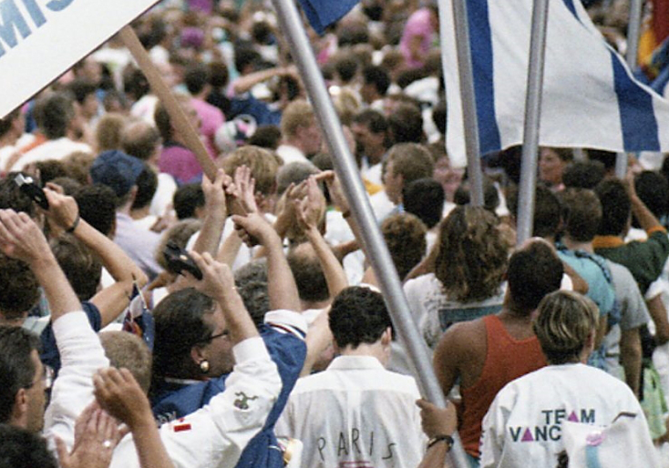 A large, tightly packed crowd of LGBTQ2S+ athletes and allies in B.C. Place Stadium, Vancouver, Canada, wave their arms and flags and sway to music at the Celebration ’90 Gay Games III Opening Ceremonies.