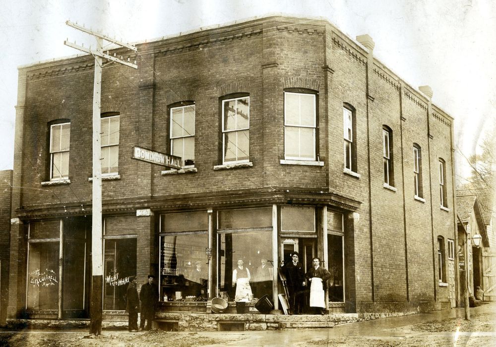 Black and white photograph of the brick exterior of a dry goods store circa 1914. Three men standing inside the shop and four men standing outside the shop. Above the shop, a sign hangs out into the street, reading 