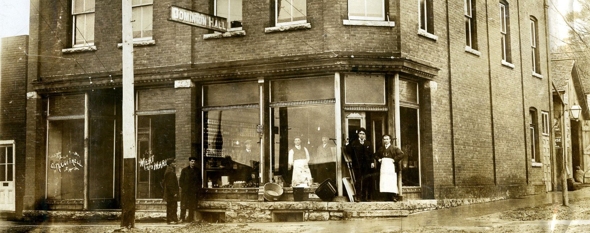 Black and white photograph of the brick exterior of a dry goods store circa 1914. Three men standing inside the shop and four men standing outside the shop. Above the shop, a sign hangs out into the street, reading 