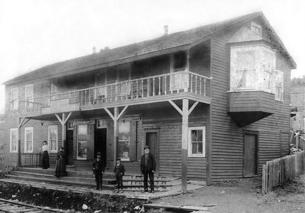 Black and white exterior. Large two storey building in background with balcony in centre and a large bay window on the right. Two women in skirts and dresses, two young boys and an older man in suits stand on the front steps of the building. There is a fence around the building and in front of the steps are railway tracks.