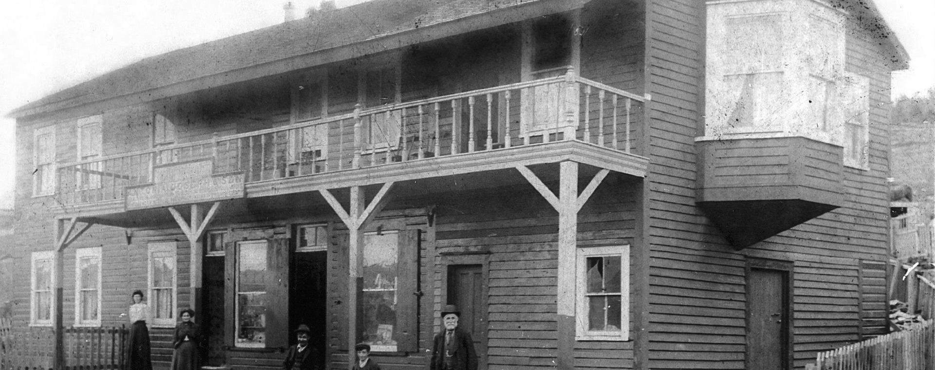 Black and white exterior. Large two storey building in background with balcony in centre and a large bay window on the right. Two women in skirts and dresses, two young boys and an older man in suits stand on the front steps of the building. There is a fence around the building and in front of the steps are railway tracks.