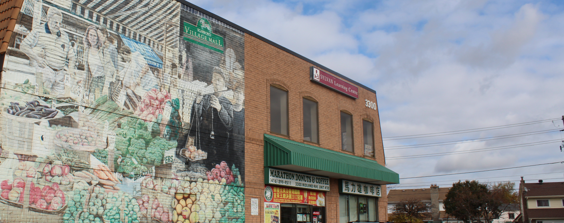 A colored photograph of a two story red brick building featuring arched windows, storefronts, and a large hand painted mural on half the building featuring a farmer's market.