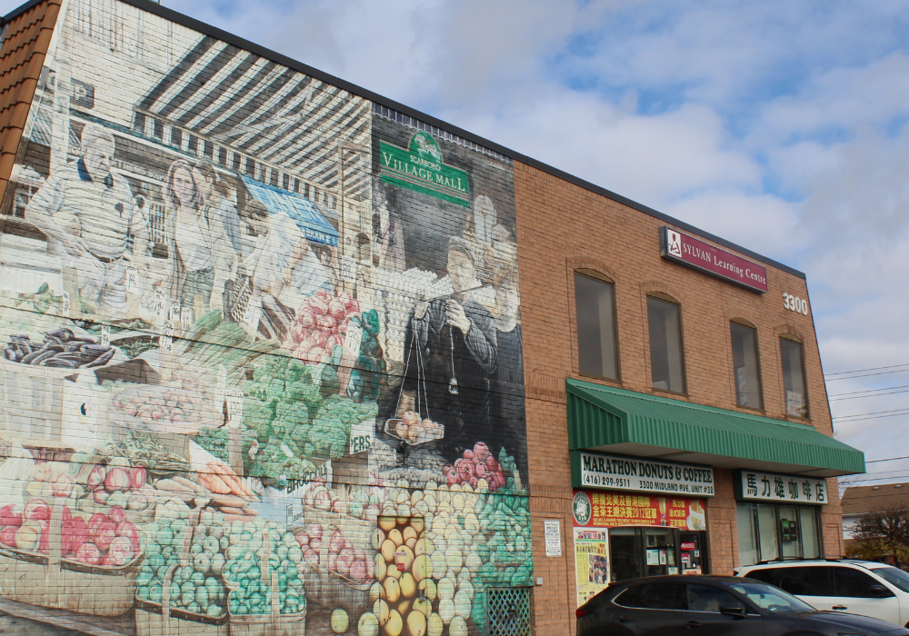 A colored photograph of a two story red brick building featuring arched windows, storefronts, and a large hand painted mural on half the building featuring a farmer's market.